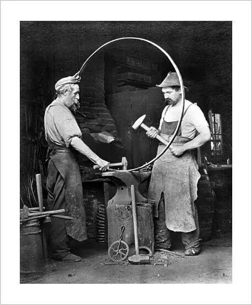 BLACKSMITHS, c1903. Two blacksmiths making a metal hoop, possibly for a wagon wheel