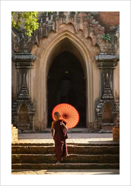 Novice Buddhist monk with red umbrella walking away from temple, Bagan, Mandalay Region