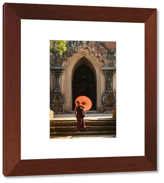 Novice Buddhist monk with red umbrella walking away from temple, Bagan, Mandalay Region