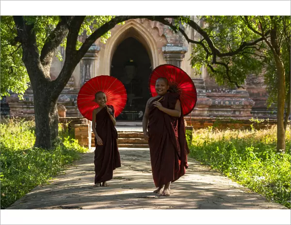 Two novice Buddhist monks with red umbrellas walking away from temple, Bagan