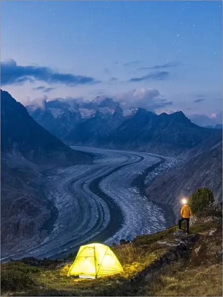 Hiker man with head torch light admiring Aletsch Glacier standing beside the tent