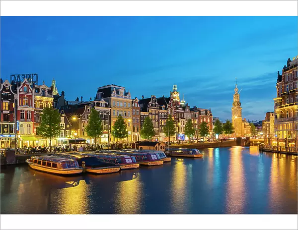 Boats moored along illuminated street on Amstel River and Munttoren tower at twilight, Amsterdam, Netherlands