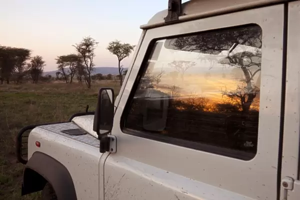 Tanzania, Serengeti. Sunrise over the bush is reflected in the window of a Land Rover