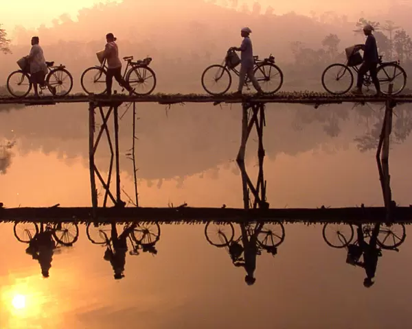 Indonesian villagers push their bicycles across a bamboo bridge as sun rises behind them outside