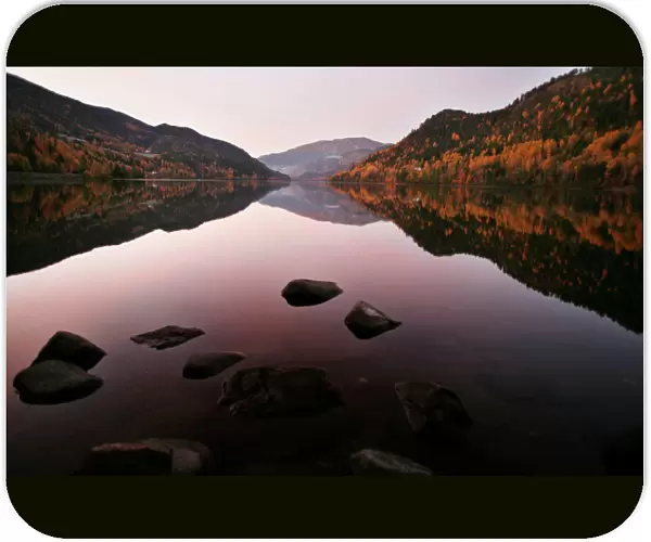 Autumn leaves are reflected in the still waters of a fjord as the sun rises west of