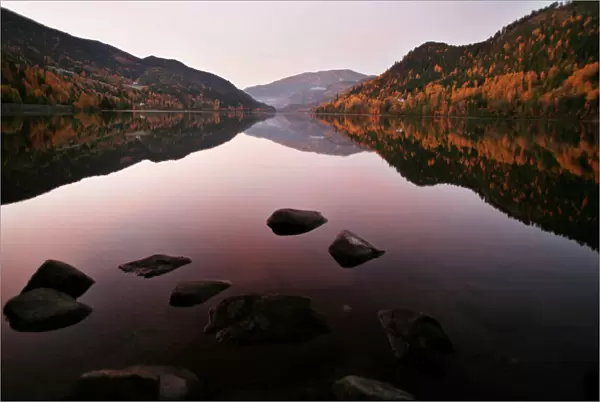 Autumn leaves are reflected in the still waters of a fjord as the sun rises west of