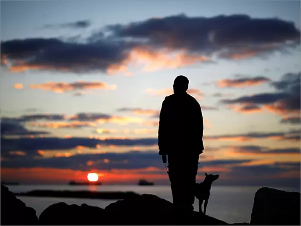 A man watches the sunset with his dog on a breakwater, along the shore of the