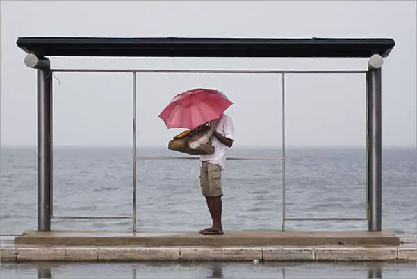 A vendor holds up an umbrella as he seeks shelter from the heavy rain under a bus stand