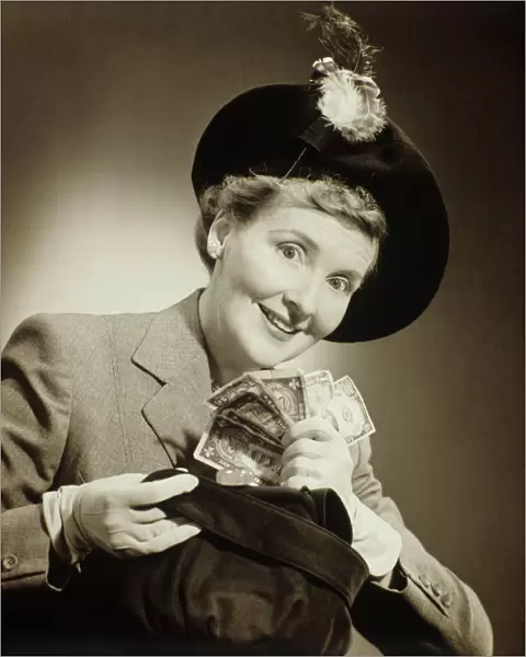 Woman in suit and fashionable hat standing taking money out of bag in studio, (B&W), portrait