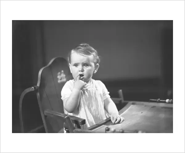 Baby girl (9-12 months) sitting in high chair, holding finger in mouth, (B&W)