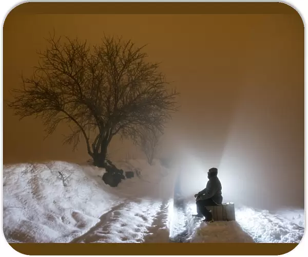Silhouette of a person sitting on a suitcase resting, for a covered way of snow and fog during the night