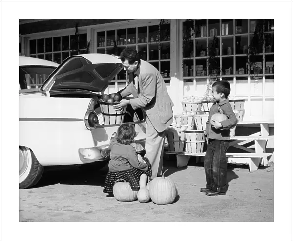 Father loading apples into trunk of car at country market, son and daughter looking