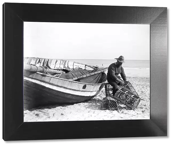 A fisherman in sou'wester mending lobster / crab creels on the beach alongside a beached Sheringham crab boat, 1910-11 (photo, glass plate negative)