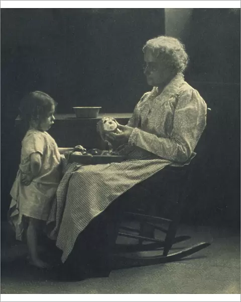 Elderly woman in a rocking chair peeling apple with young girl standing in front of her, c1900. Creator: Elizabeth B. Brownell