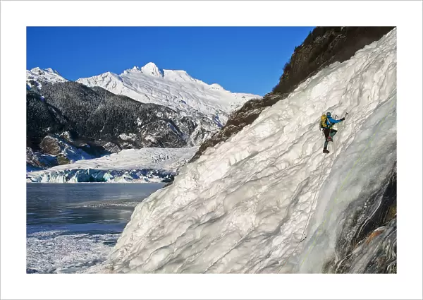 Female Ice Climber Climbs The Frozen Nugget Falls With Mendenhall Glacier In The Background, Juneau, Southeast Alaska, Winter