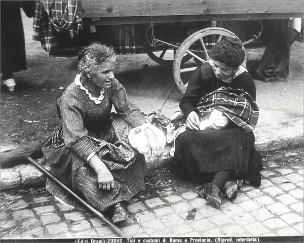 An elderly woman and a young woman seated on a sidewalk in Rome, in front of a cart