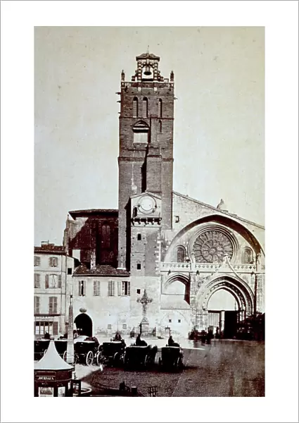 The facade of the Church of St-Etienne in Toulouse. In the foreground place St-Etienne with a few parked gigs, a small ornamental obelisk and a newstand