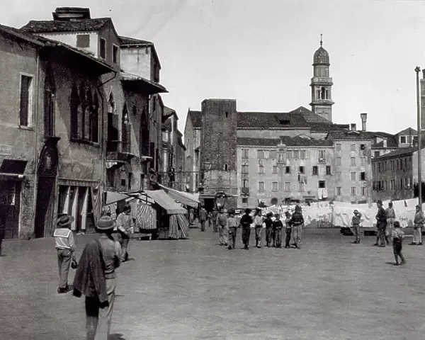 View of Campo Margarita in Venice, crowded with pedestrians. On the left, a few stands. In the background a row of drying laundry. Two bell towers stand out from between the roofs of the houses