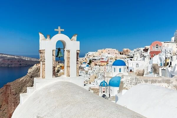 A church roof and bell with the white washed stone walls and blue church cupolas of Oia