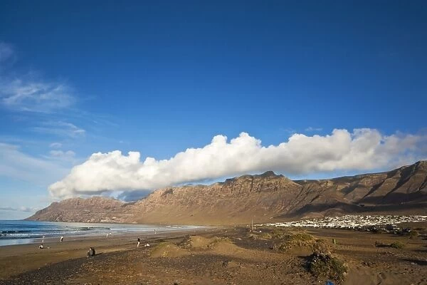 Spectacular 600m volcanic cliffs of the Risco de Famara rising over Lanzarotes finest beach at Famara, with its low-rise bungalow development, Famara, Lanzarote, Canary Islands, Spain, Atlantic