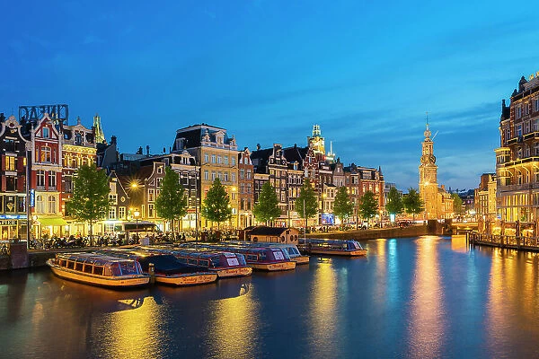 Boats moored along illuminated street on Amstel River and Munttoren tower at twilight, Amsterdam, Netherlands