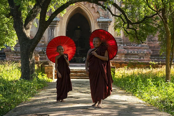 Two novice Buddhist monks with red umbrellas walking away from temple, Bagan