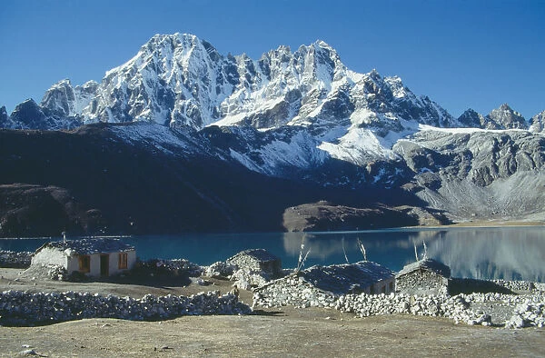 Nepal, Himalayas, Gokyo, Snow covered peaks overlooking lake with stone built houses and livestock enclosures