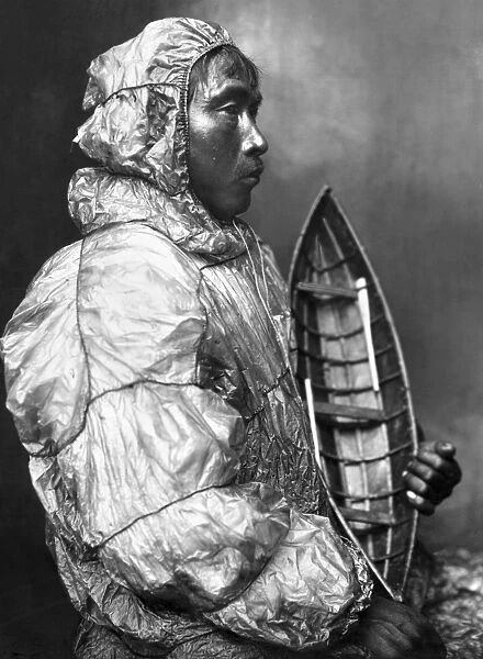 An Eskimo kayaker wearing a waterproof jacket and holding a toy boat he made for his son. Photgraphed by the Lomen Brothers, early 20th century