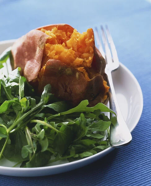Baked sweet potato served with rocket leaves, on white plate with silver fork, standing on blue tablecloth