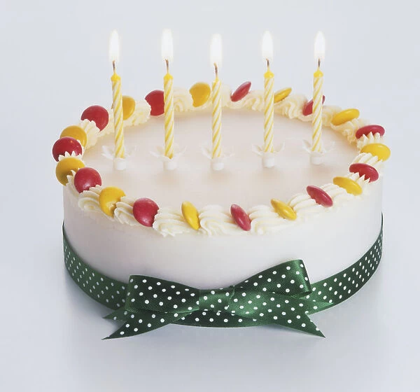 Birthday cake decorated with five candles, white icing, red and yellow sweets, and green ribbon tied around it