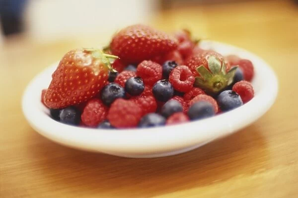 Blueberries, strawberries and red raspberries in round white dish on wooden table