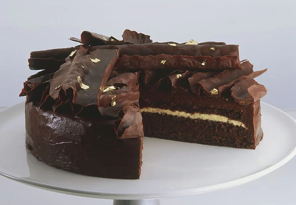 Chocolate cake with section removed on cake stand, filled, iced, decorated with chocolate curls and gold leaf, close up