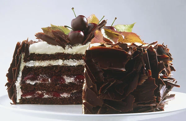 Chocolate cake topped with white cream, fresh cherries and autumn leaves, sides coated with chocolate curls and section removed to reveal cross-section, side view