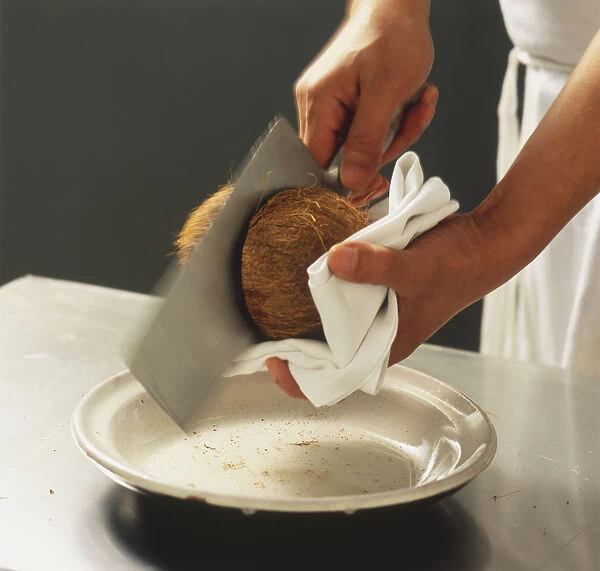 Cocos nucifera, Coconut, being sliced in half with large blade, one hand holding coconut and other hand cutting, ceramic plate underneath