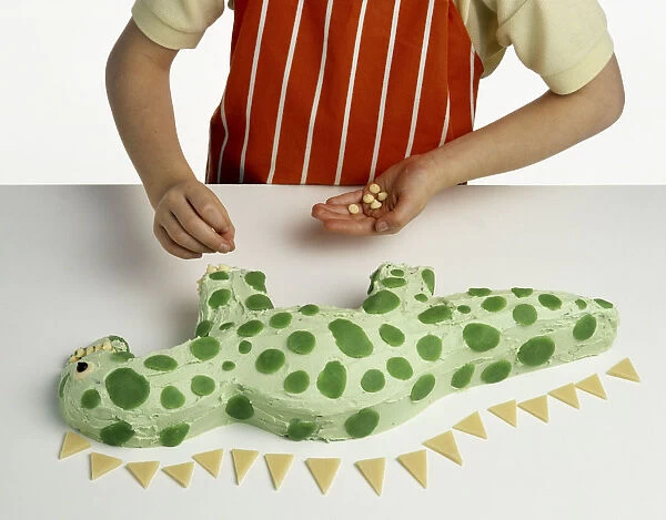 Hand model wearing orange and white striped apron, decorating green dinosaur-shaped cake with white chocolate buttons