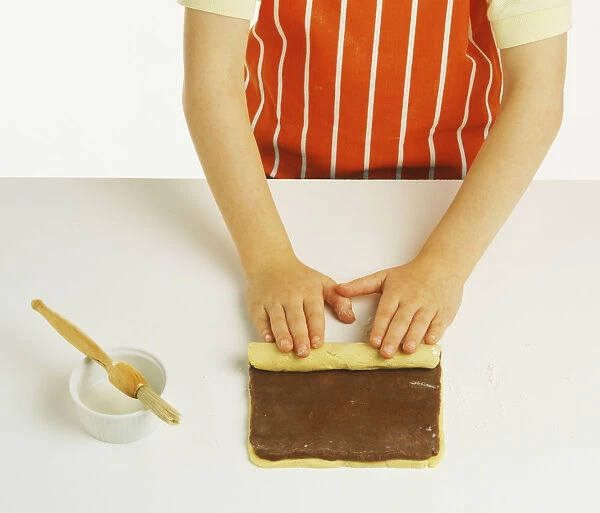 Hand model wearing an orange and white striped apron, rolling together flattened plain and chocolate pastry