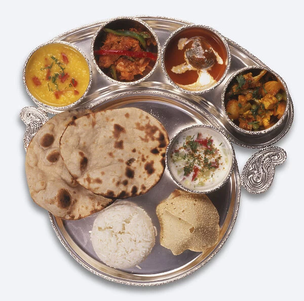Indian Thali served on a silver platter, containing large round Roti, white Rice; crispy Papad, white Raita, Mixed vegetables, red and white Malai kofta, Kadhai murg, yellow coloured Dal, above view