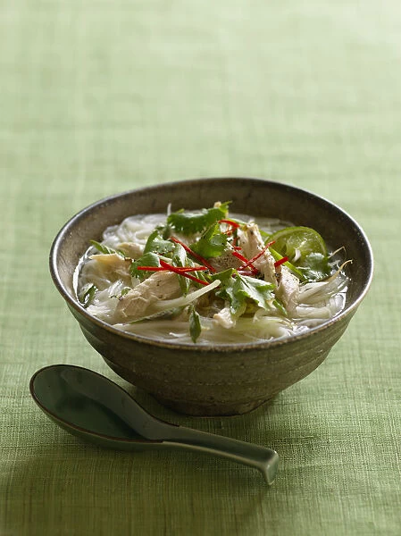 Pho ga, bowl of Vietnamese rice noodle soup with chicken, garnished with herbs, red chilli and lime, with spoon nearby