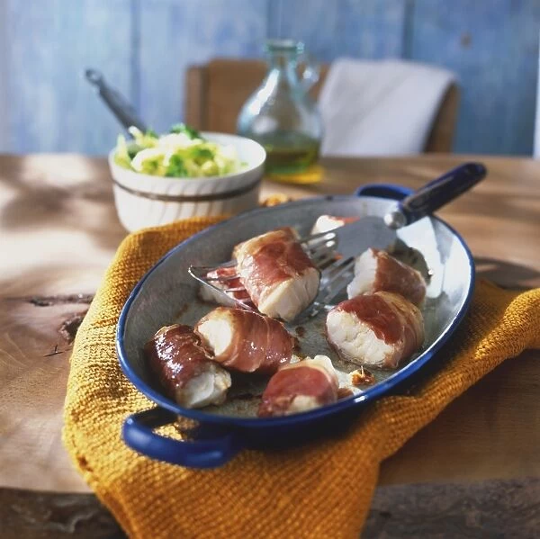 Pieces of cod wrapped in parma ham, in blue pan, spatula embedded, bowl of green salad and bottle of olive oil in background, on wooden table