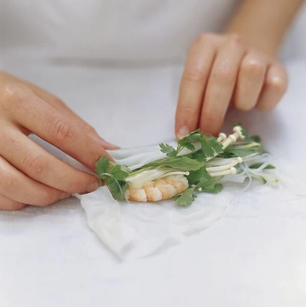 Rolling up rice paper wrapper stuffed with enoki mushrooms, prawns and herbs, close-up