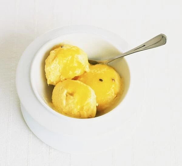 Three scoops of ice cream topped with mango syrup served in white bowl with spoon, view from above