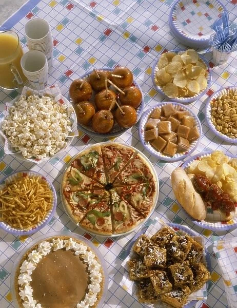 Selection of party food dishes on a table, including pizza slices, cakes, toffee apples, crisps and popcorn on paper plates, orange juice and paper cups