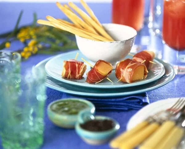Table laid with Mediterranean appetizers, bacon-wrapped fruit cubes on cocktail sticks, bowl of bread sticks, bowls of dips