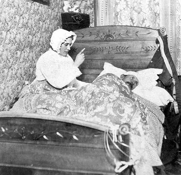Bedding scene: a couple fighting; they both wear a night cap. 1900 circa Historic photograph, woman scolding her husband in bed, around 1900