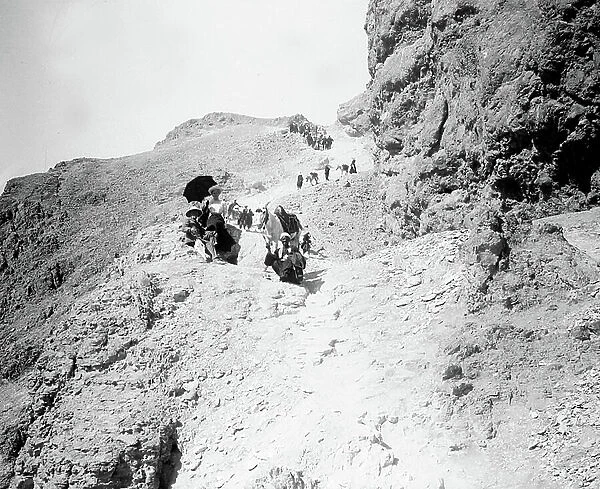 Egypt: Cook cruise, trip in the valley of the Kings amidst stones and dust, group of tourists with guides and mules. Tourists with umbrella and colonial hat, 1900