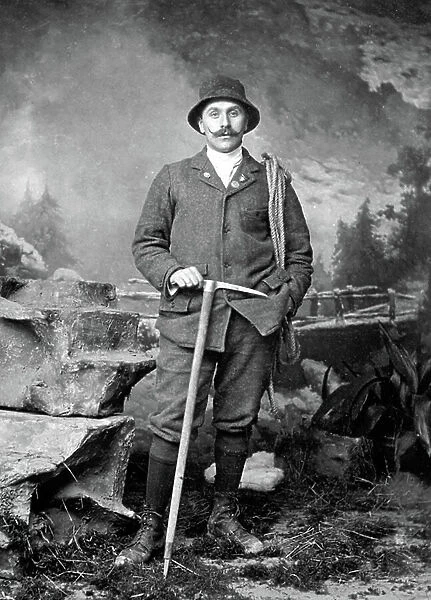 Full-length portrait of a man dressed as a mountain climber. He is leaning on a pick and has a rolled up rope on his left shoulder. Behind him a mountain backdrop