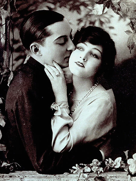 Half-length portrait of a young couple leaning against a balcony decorated with plants. The man has his arm around the girl's waist and is kissing her cheek