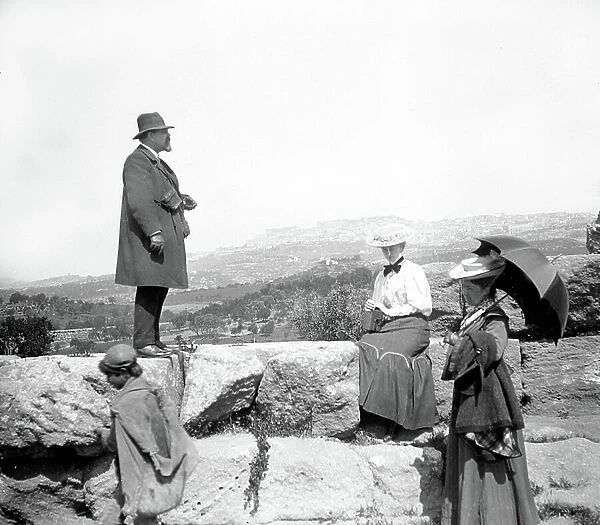 Italy, Sicily: tourists with umbrella and camera visit Roman ruins at the foot of a big city, 1900