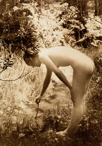 A young woman, nude in the woods, seen in profile as she bends over to put a branch in a stream
