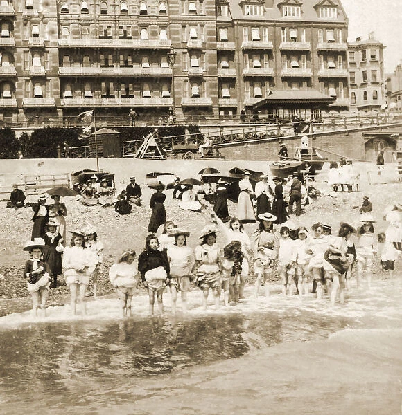 Historic image in sepia of a group of children standing in a row in shallow water with their skirts and knickers pulled up, adults sitting on the beach; Brighton, England